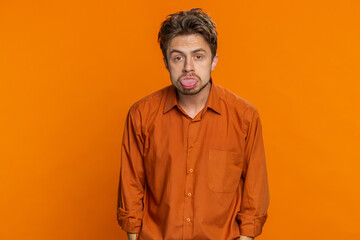 Funny Caucasian man showing tongue making faces at camera, fooling around, joking, aping with silly face, teasing, bullying, abuse disrespect. Handsome young guy isolated on orange background indoors