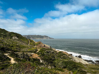 California coast with views on the waters of the bay on the bright day. Bushes and flowers of the West Coast nature in front of the lightly cloud skies. Waves of the Pacific Ocean waters of the bay.