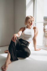 A blonde woman in a white crop top and black pants sits by a window, bathed in natural light. She exudes confidence and style...