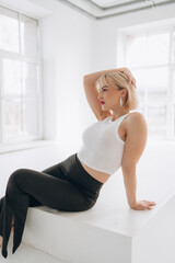 A blonde woman with red lipstick in a white crop top and black pants poses on white steps in a bright, minimalist room. She leans back gracefully.