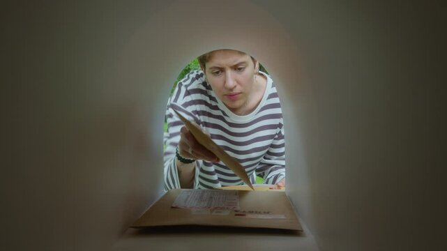 Young woman retrieving envelopes from mailbox and checking information on postal labels. View from inside of mailbox