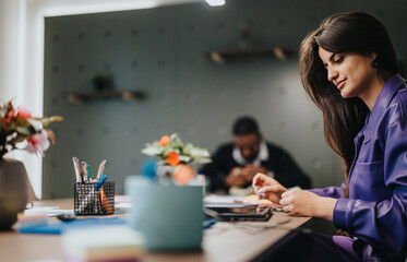 Professional woman in a purple blazer focusing intently on her work at a modern office desk, with colleagues in the background.