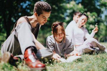 Fototapeta premium A group of young friends relax together in a park, examining nature up close, sharing a delightful moment under the shade of trees with vibrant greenery around.