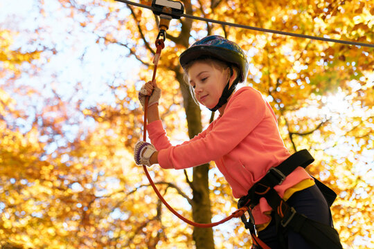 Kid clambering in adventure park with protective helmet and safety tourist equipment. Child girl climbing on rope obstacle courses at nature. Outdoor sports camp in autumn forest. Healthy lifestyle