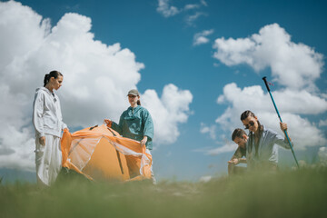 Four friends set up an orange tent in a scenic outdoor area with a bright sky and clouds, enjoying...