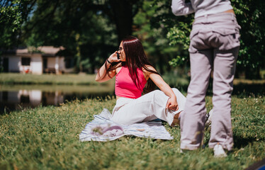 Obraz premium A joyful young woman in bright casual wear relaxes on a picnic blanket in a lush green park, chatting with a friend on a sunny day.