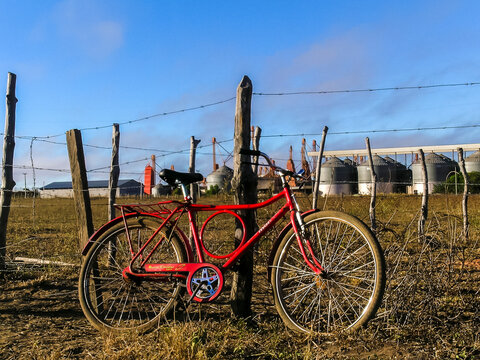 Luis Eduardo Magalhaes, Bahia, Brazil, June 16, 2004. Red bicycle from the brazilian brand Monark stopped next to a barbed wire fence with grain silos from a farm in the background
