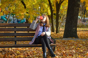 a girl sits on a bench in an autumn park, headphones on her head, a cup of drink in her hands: tea, coffee or cappuccino. child listens to music or audiobook. hobbies, recreation and entertainment