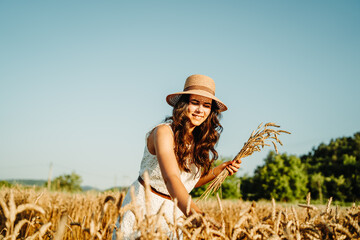 One beautiful young caucasian woman wearing white dress and straw hat holding crops of wheat and standing in wheat field during the day 