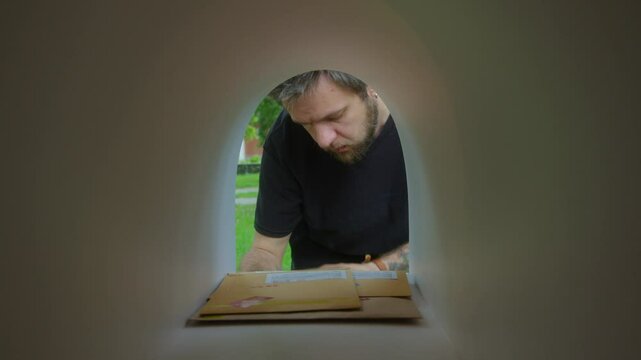 Male recipient picking up parcel and envelopes from front yard mailbox and reading information on labels. View from inside of mailbox