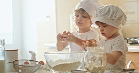 Two toddlers in chef hats are cooking in a kitchen, sharing tableware