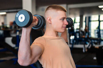 A young bodybuilder lifts dumbbells while seated on a bench in a gym