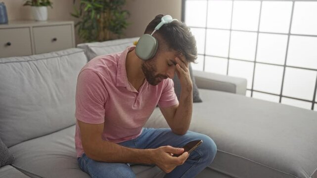 Young man wearing headphones and pink shirt sits on a gray sofa in a modern living room, looking at his phone thoughtfully with a hand on his forehead, surrounded by indoor plants