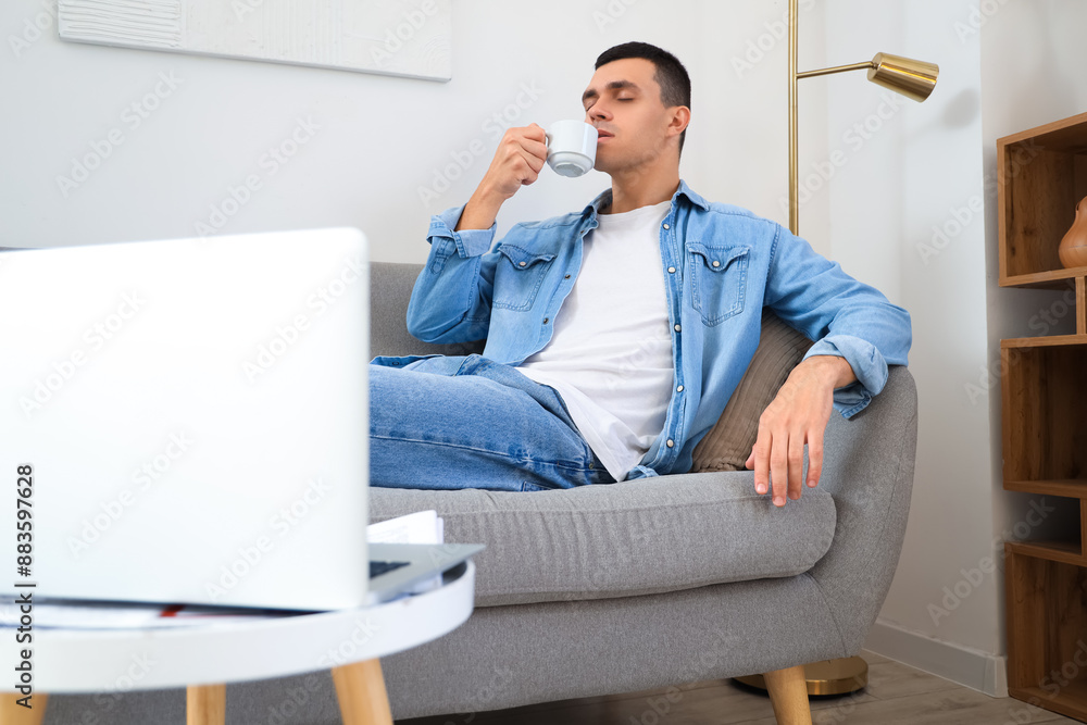 Poster young man drinking espresso while lying on sofa with laptop on coffee table