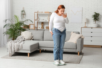 Shocked overweight woman in tight jeans standing on scales at home. Excess weight concept