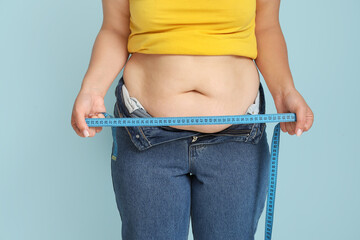 Overweight woman in tight jeans measuring her belly on blue background. Excess weight concept