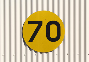 Round yellow sign with the number 70 in black, mounted on a corrugated metal wall