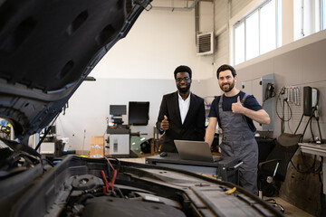Open car hood in foreground and laptop on tool cart. Mechanic and businessman giving thumbs up in car repair shop. Concept of car repair, teamwork, and customer satisfaction.