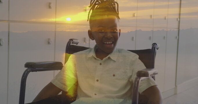 Image of sunset landscape over happy african american schoolboy in wheelchair by lockers