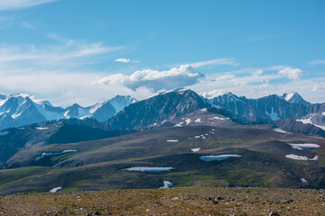Top view from sunlit grassy stony plain of pass to high snow mountains in bright sun under clouds...