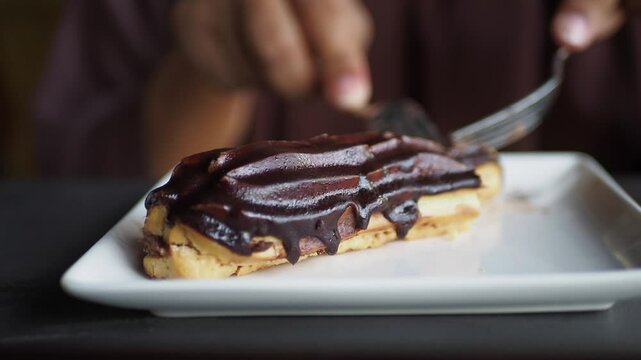 In the image, someone is using a fork and knife to slice a chocolate eclair on a plate