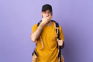 Caucasian handsome man with backpack and trekking poles over isolated background covering mouth with hand