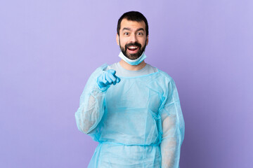 Surgeon man with beard with blue uniform over isolated purple background surprised and pointing front