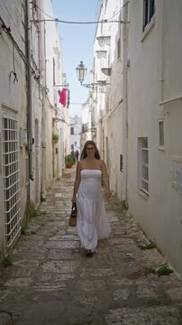 A young hispanic woman in a white dress walks through the old town streets of ostuni in puglia, italy on a sunny day, capturing the charming european architecture.