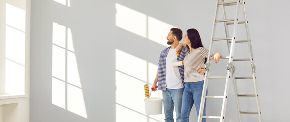 Family couple doing renovations at home. Young man and woman change color of walls in living room. Banner with happy husband and wife standing by ladder and looking at freshly painted light gray wall © Studio Romantic
