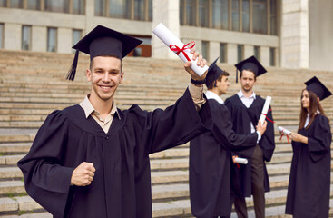 Happy male student enjoying his graduation day. Portrait of joyful young man in cap and gown standing outside college or university building, holding up his diploma, looking at camera and smiling