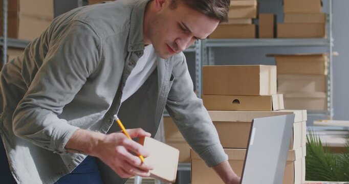 Serious, attentive male worker in modern warehouse prepares order for shipment. Man takes notes during process of labeling and arranging packages, focusing on detail accuracy for e-commerce.