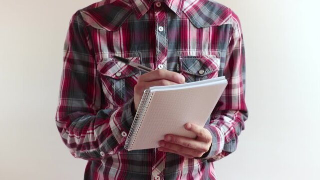 man with checkered shirt takes notes, he's against a white background
