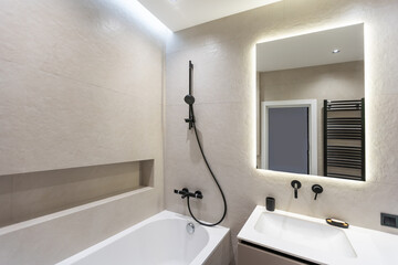 Bathroom in a modern style with gray and white tiles. There is a large mirror with luminous lamps, tabletop with wooden drawers and sink, bath with shower and glass partition, towel rack and a hanger.