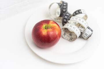 Slimming. A red-orange apple wrapped in a measuring tape lies on a white plate, on a white background. Calls to lose weight