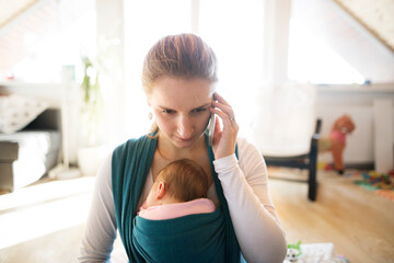 Newborn baby sleeping in baby sling, wrap, mother making phone call on smartphone. Unconditional...