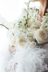 The morning of the bride, a woman wearing a white silk robe holds a wedding bouquet. Close-up photo.