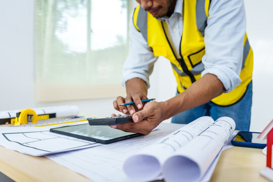 Asian male architect works at desk with house model toy and A3 paper blueprints, construction real estate law, zoning, permits, and resolving property disputes through arbitration and litigation.