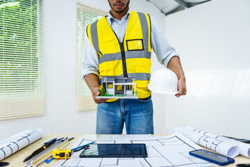Asian male architect works at desk with house model toy and A3 paper blueprints, construction real...