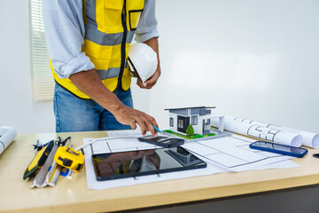 Asian male architect works at desk with house model toy and A3 paper blueprints, construction real...