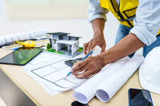 Asian male architect works at desk with house model toy and A3 paper blueprints, construction real estate law, zoning, permits, and resolving property disputes through arbitration and litigation.