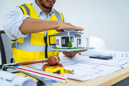 Asian male architect works at desk with house model toy and A3 paper blueprints, construction real estate law, zoning, permits, and resolving property disputes through arbitration and litigation.