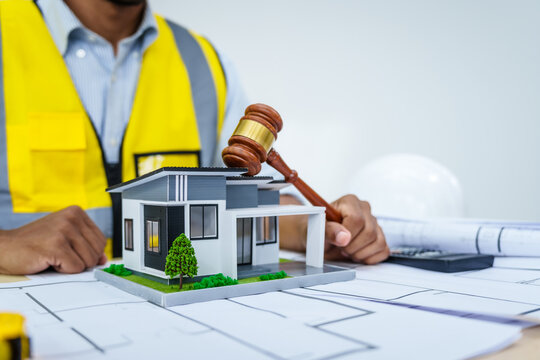 Asian male architect works at desk with house model toy and A3 paper blueprints, construction real estate law, zoning, permits, and resolving property disputes through arbitration and litigation.
