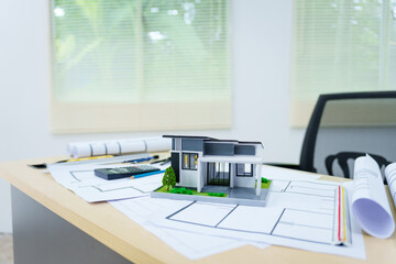 An architect's desk with no people, featuring a modern house model toy and A3 paper blueprints, set against a modern indoor workspace.