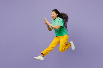 Full body young woman of African American ethnicity with dreadlocks in green t-shirt casual clothes jump high hold in hand use mobile cell phone isolated on plain purple background Lifestyle concept