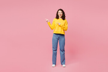 Naklejka premium Full body smiling happy young woman wearing yellow sweatshirt casual clothes point index finger aside on area mock up isolated on plain pastel light pink background studio portrait. Lifestyle concept.
