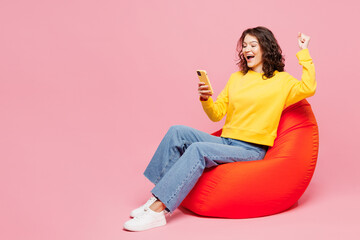 Full body young woman wearing yellow sweatshirt casual clothes sit in bag chair hold in hand use...