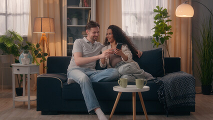 Fototapeta premium Multiracial positive happy couple family Caucasian man guy male Hispanic Latino woman girl female smile laughing talking using mobile phone together funny video watching shopping online at couch home