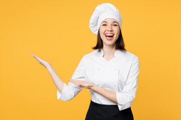 Young surprised chef cook baker woman she wears white shirt black apron uniform toque chefs hat point hands arms aside on area mock up isolated on plain yellow background studio. Cooking food concept.