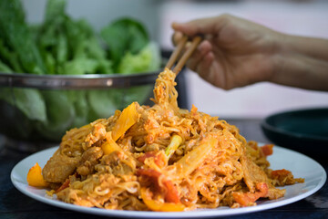 A hand use chopsticks  pick up stir fried instant noodles in white plate on the table and fresh green vegetable in basket. Selective focus with blur background closed up.