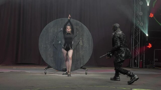 A circus show with throwing knives in the circus arena under the light of colorful spotlights. The assistant girl stands near the wall where a man with a mask on his face throws knives.
